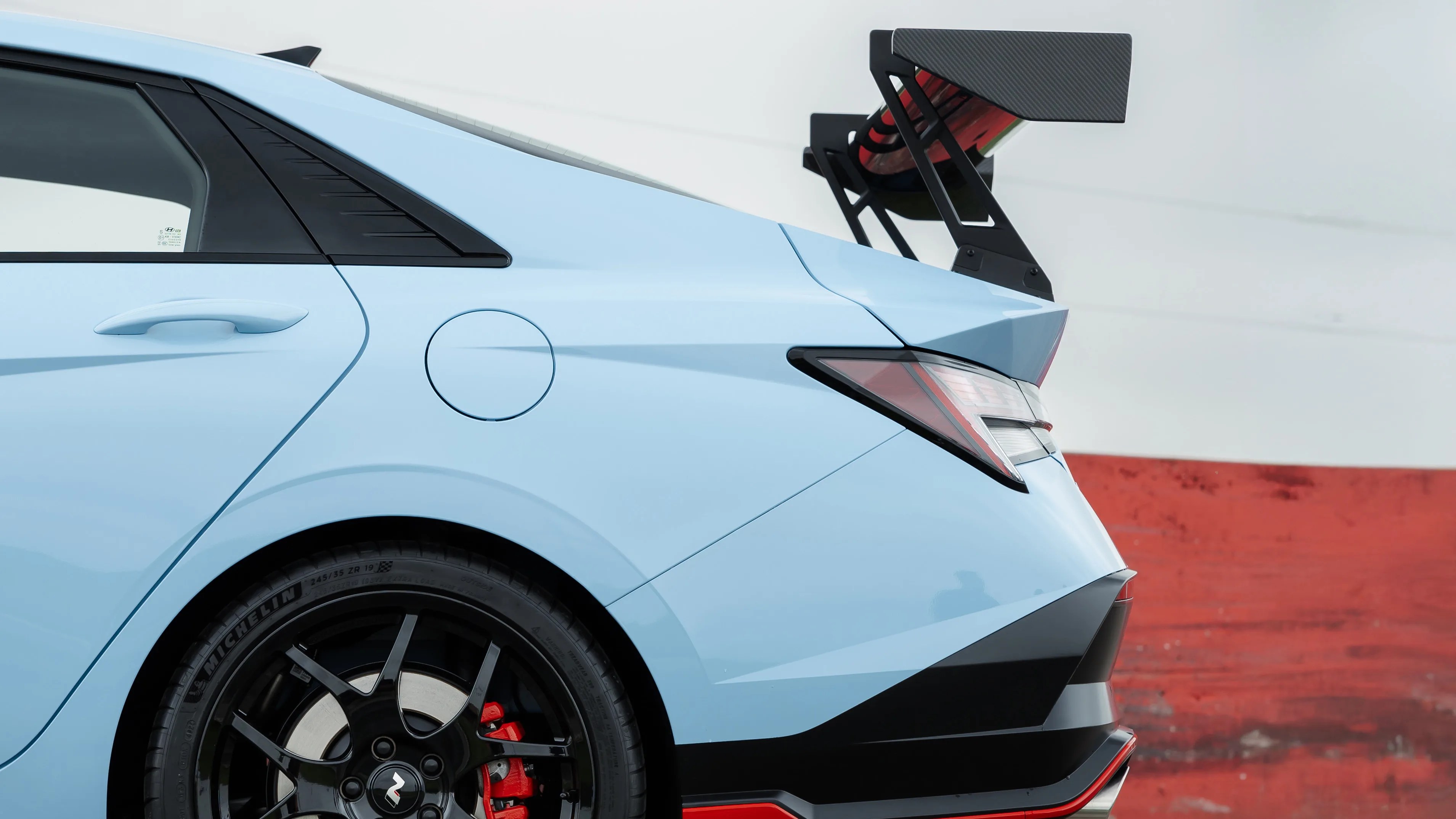Light blue sports car rear side with black alloy wheel, red brake caliper, and large carbon fiber rear wing.