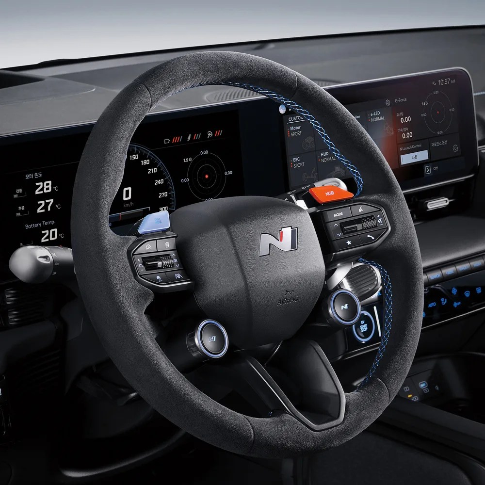Black suede steering wheel with blue stitching and N logo in a car interior with digital displays.