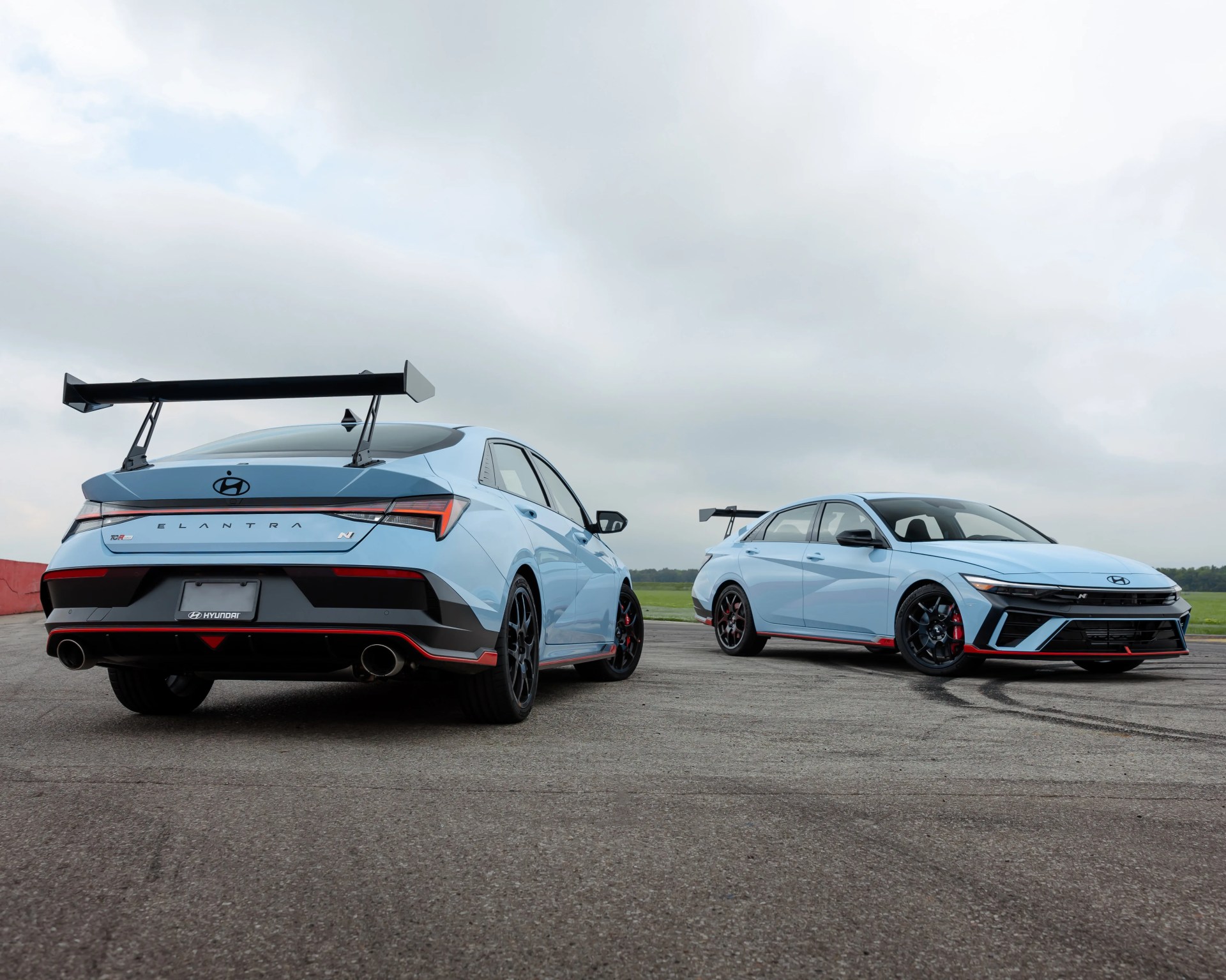 Two light blue Hyundai Elantra N sports sedans with black rear wings parked on an asphalt surface under a cloudy sky.