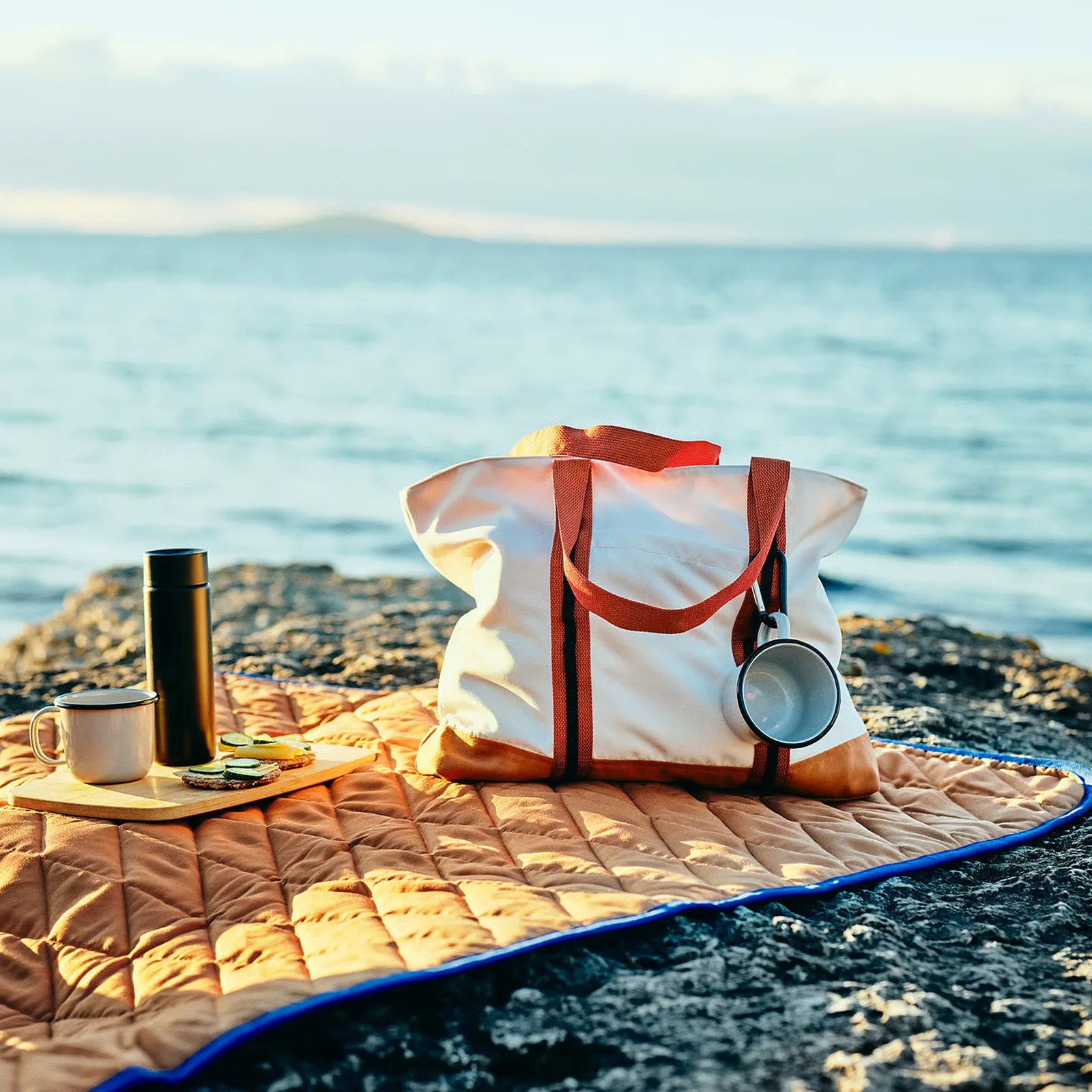 White and brown canvas tote bag with a metal cup attached, placed on a brown quilted blanket by the sea.