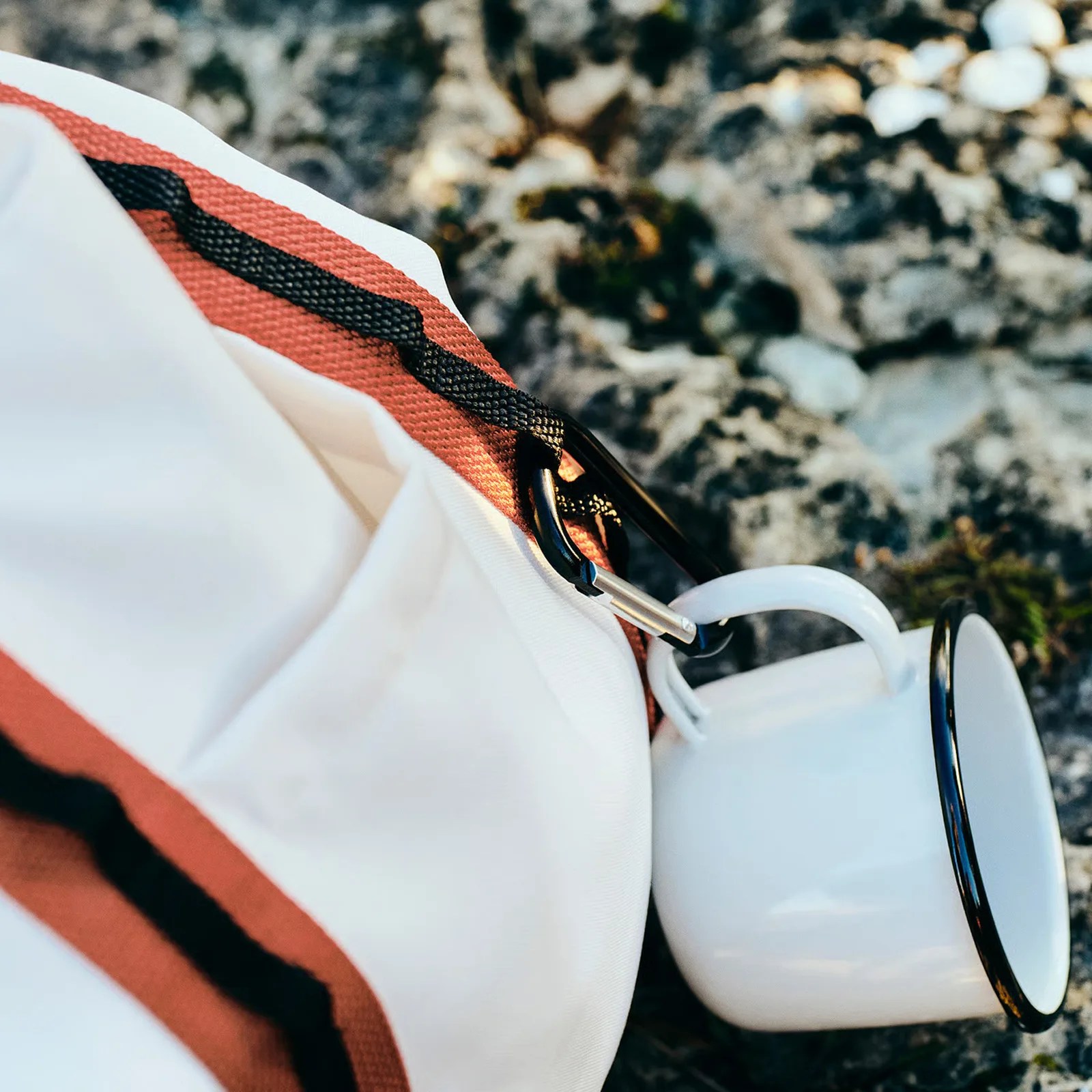 White enamel camping mug with black rim attached to a white and orange fabric bag by a black carabiner.