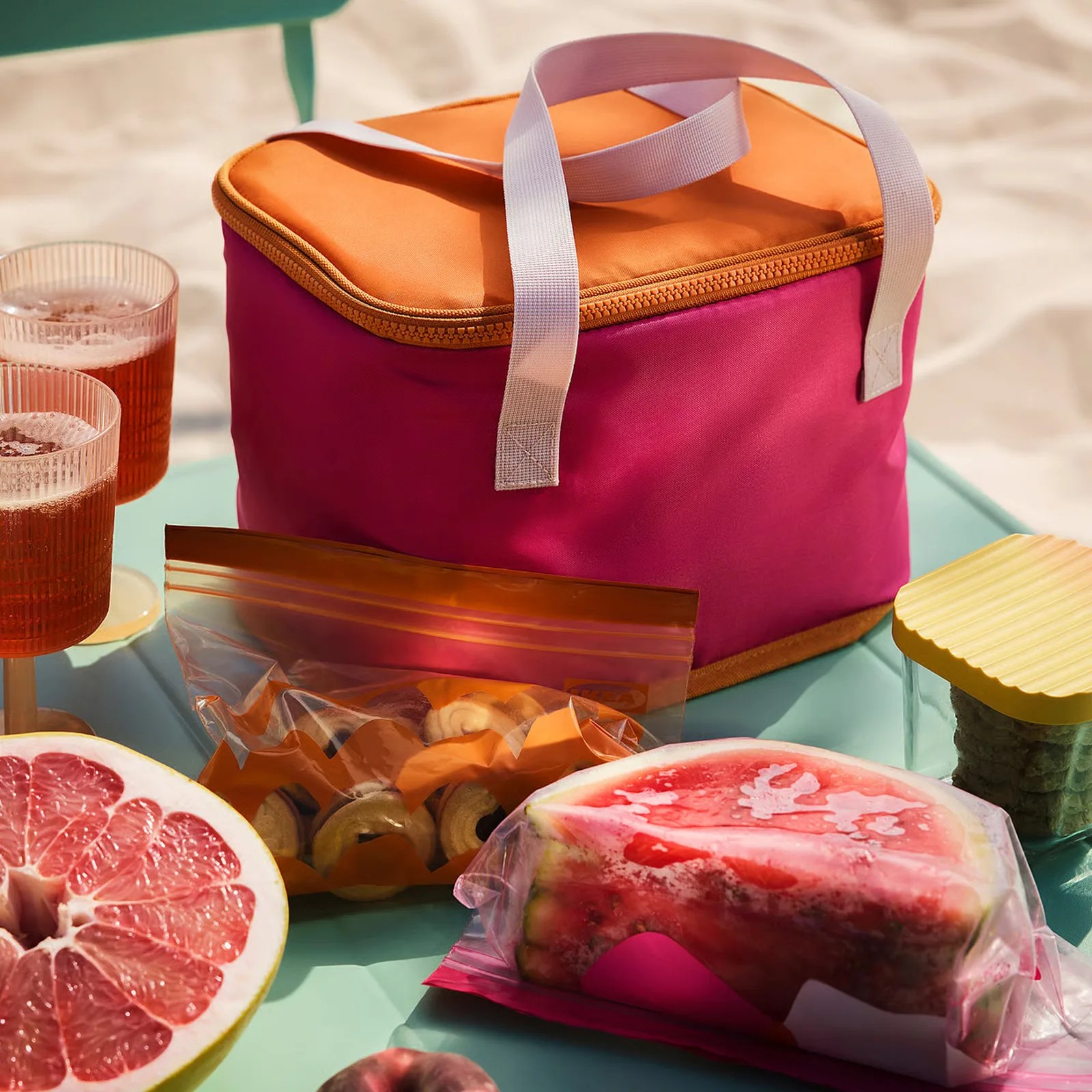 Pink and orange soft-sided cooler bag with white handles on a table with snacks and drinks.