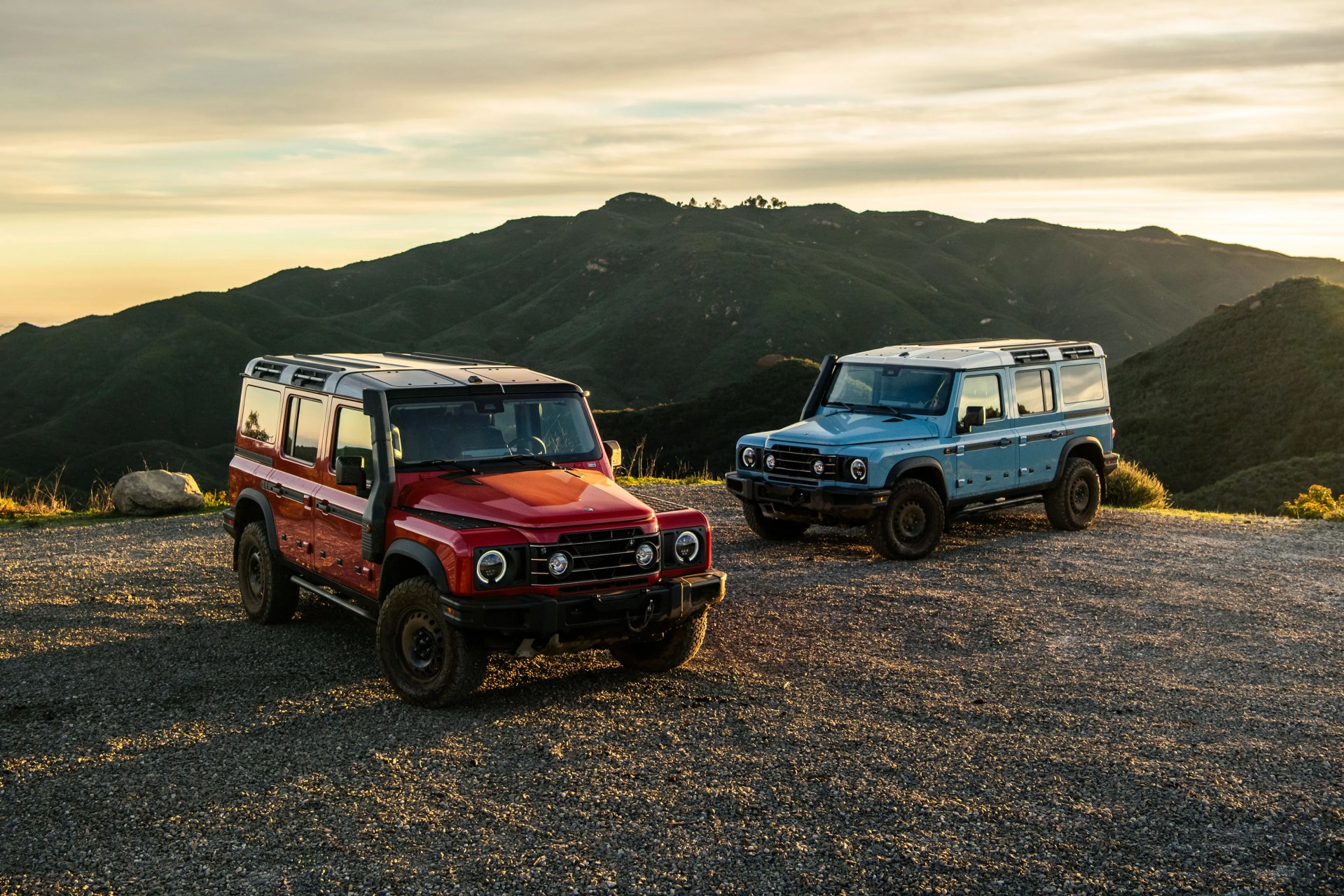 Two off-road SUVs, one red and one light blue, parked on a gravel area with green mountains in the background at sunset.
