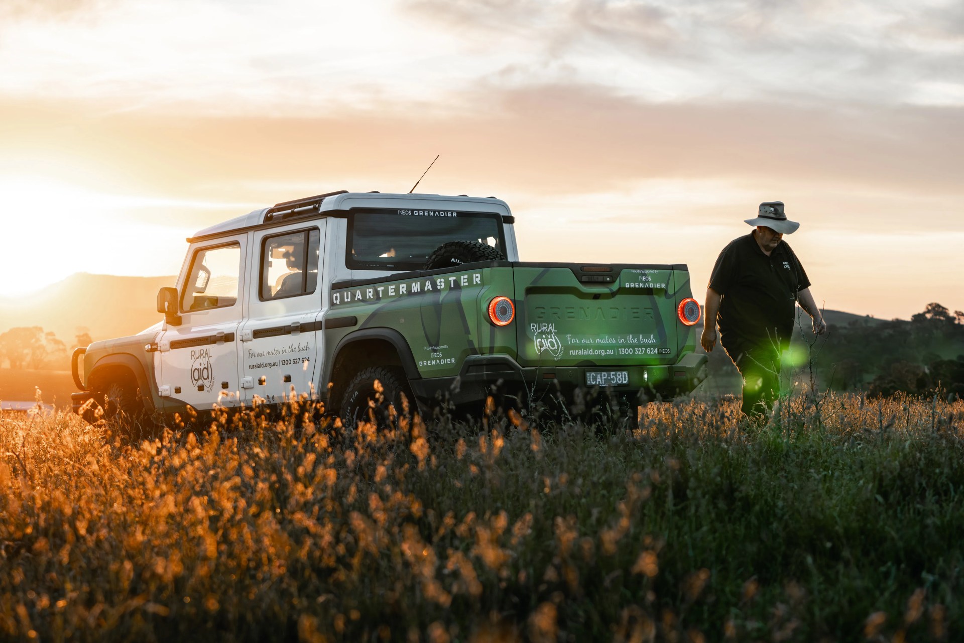 White and green Ineos Grenadier Quartermaster pickup truck parked in a field at sunset with a person wearing a hat walking nearby.