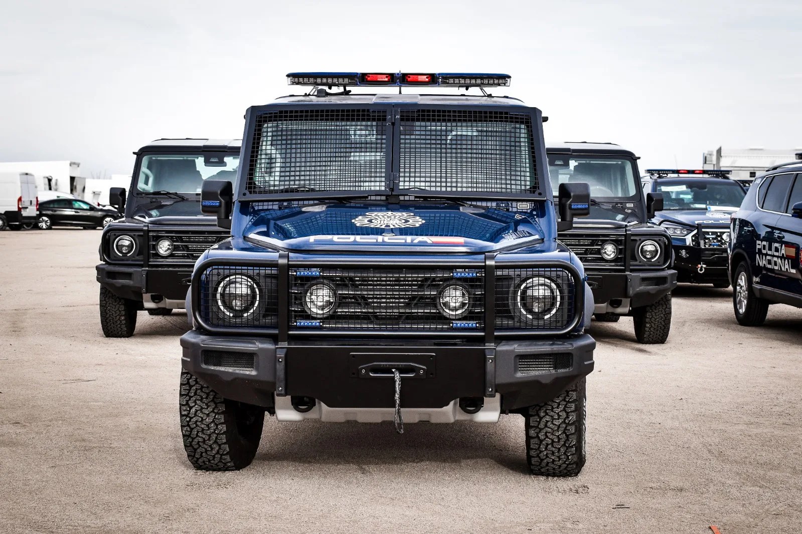 Blue police armored vehicle with protective grille and lights, flanked by similar vehicles in a parking lot.