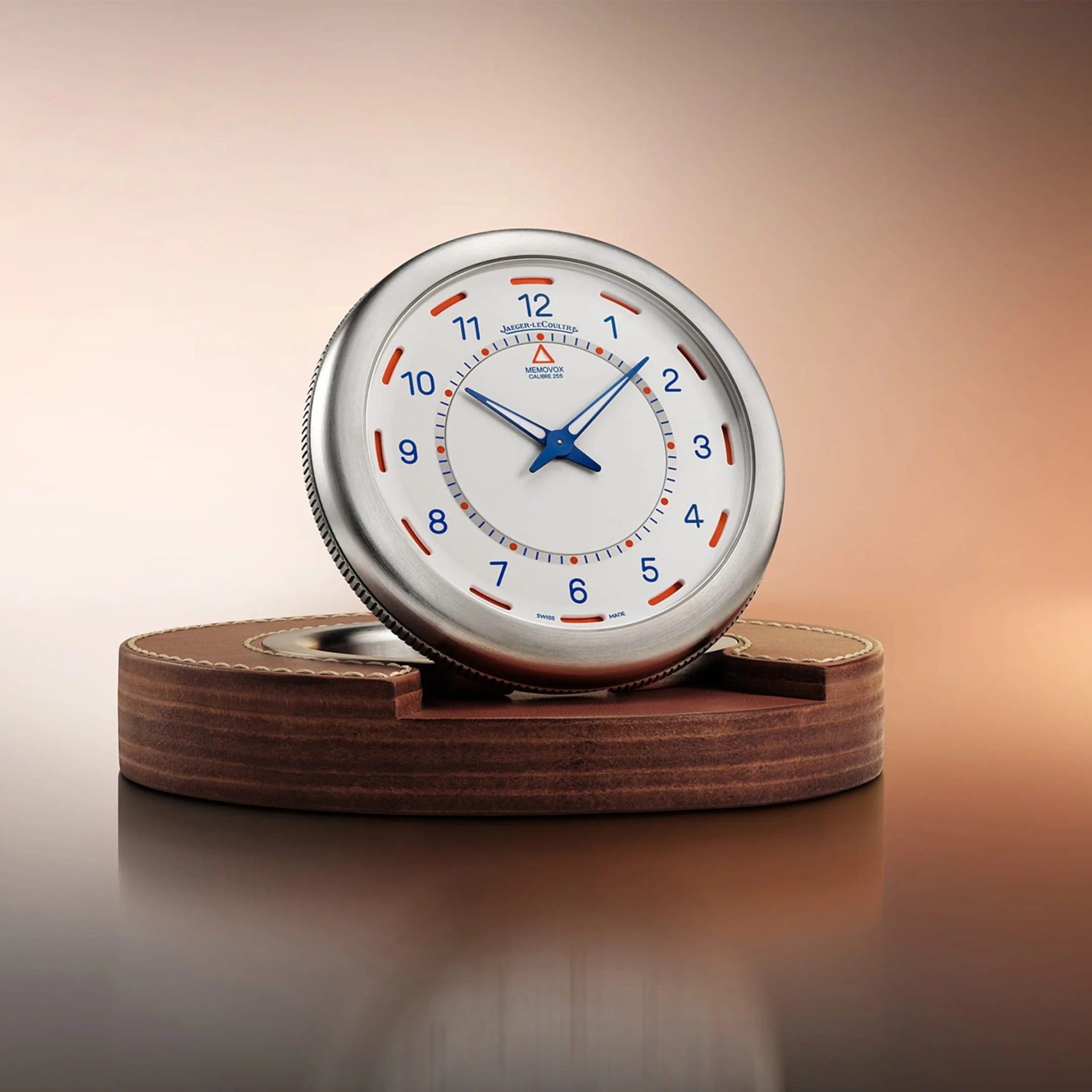 Round silver Jaeger-LeCoultre Memovox clock with blue hands and numerals on a brown leather stand.