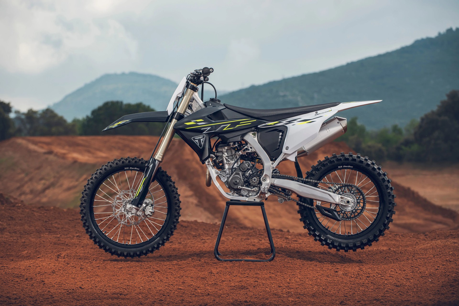 Black and white Triumph dirt bike with yellow accents on a dirt track with hills in the background.