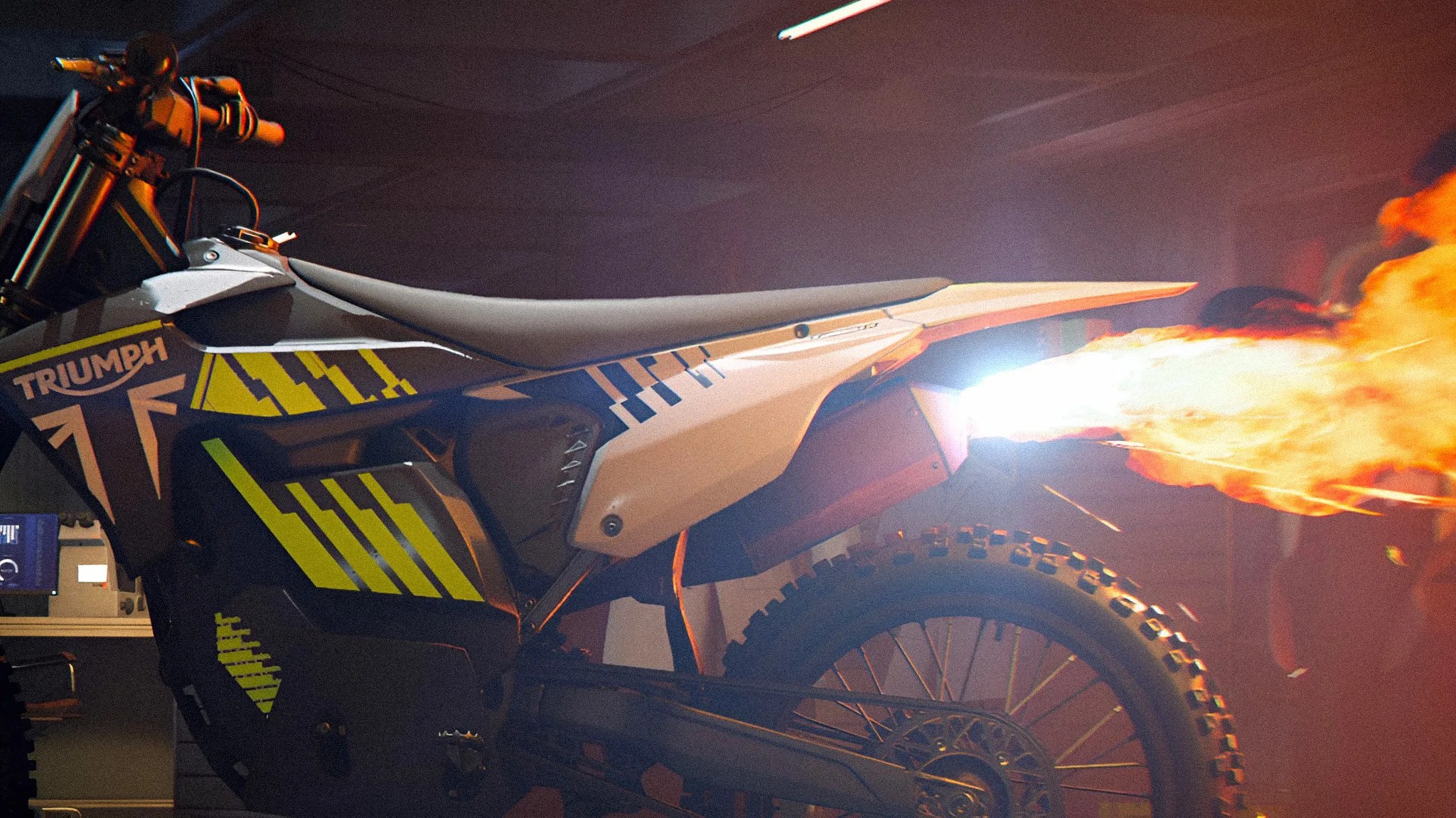 White and black Triumph dirt bike with yellow graphics emitting flames from the exhaust in a dimly lit garage.