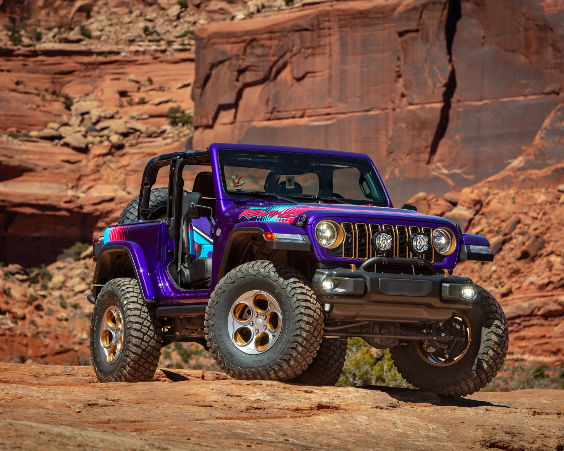 Purple off-road Jeep Wrangler with large tires and colorful decals parked on rocky terrain with red rock cliffs in the background.