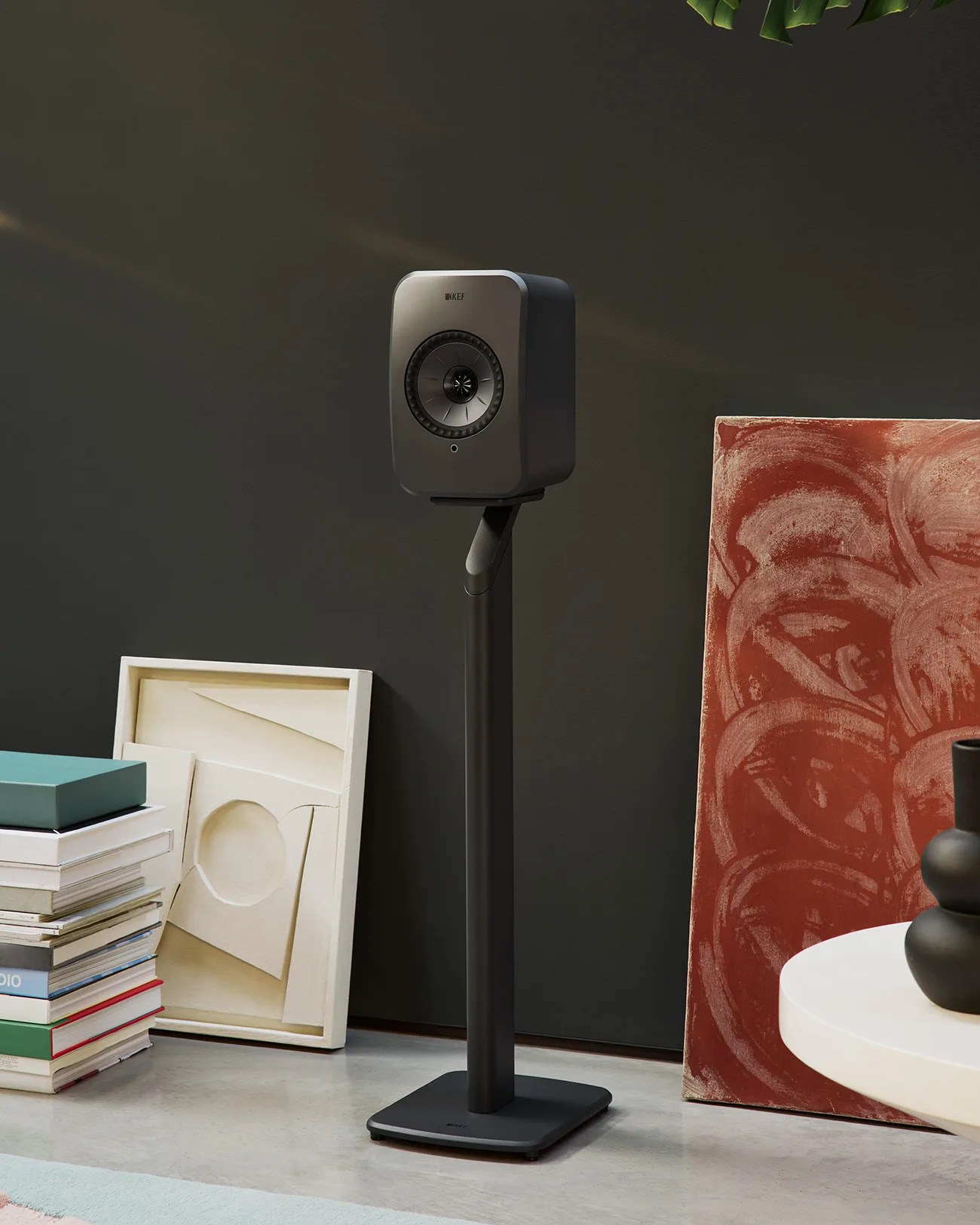 Black KEF speaker on a tall black stand against a dark wall with stacked books and abstract art nearby.