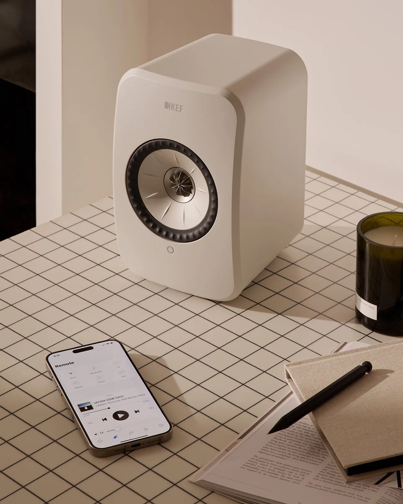 White KEF speaker on a grid-patterned table next to a smartphone, a black pen, a beige notebook, and a dark green candle.