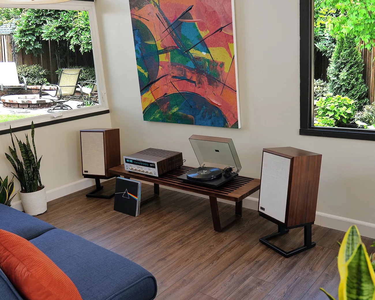 Wooden audio setup with vintage speakers, turntable, and amplifier on a slatted bench in a living room with large windows.