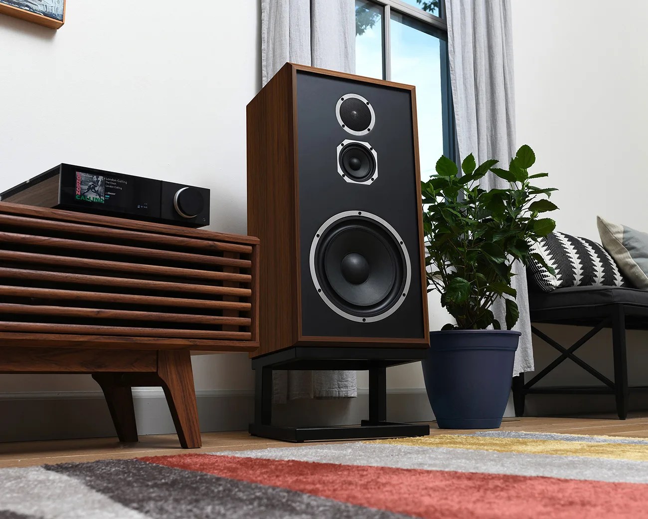 Large vintage-style wooden speaker with three drivers on a black stand next to a potted plant and wooden cabinet.
