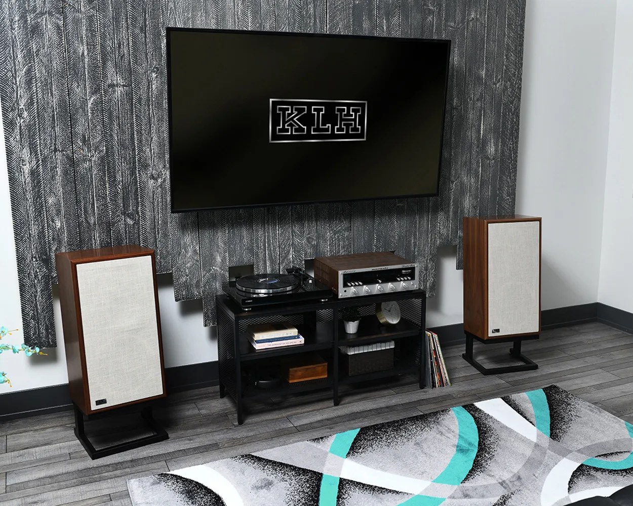 Living room audio setup with two wooden KLH speakers with beige fabric covers on black stands, a black metal shelf holding a turntable and amplifier, and a wall-mounted TV displaying the KLH logo.