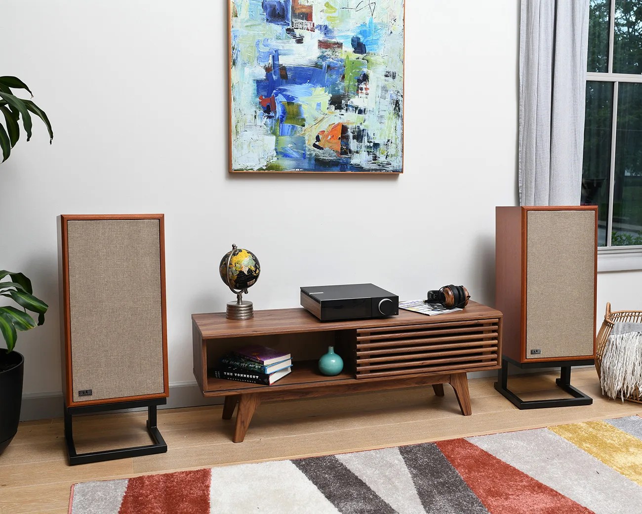 Mid-century modern wooden media console with slatted door, black stereo receiver, globe, books, and headphones, flanked by two vintage speakers on black metal stands.