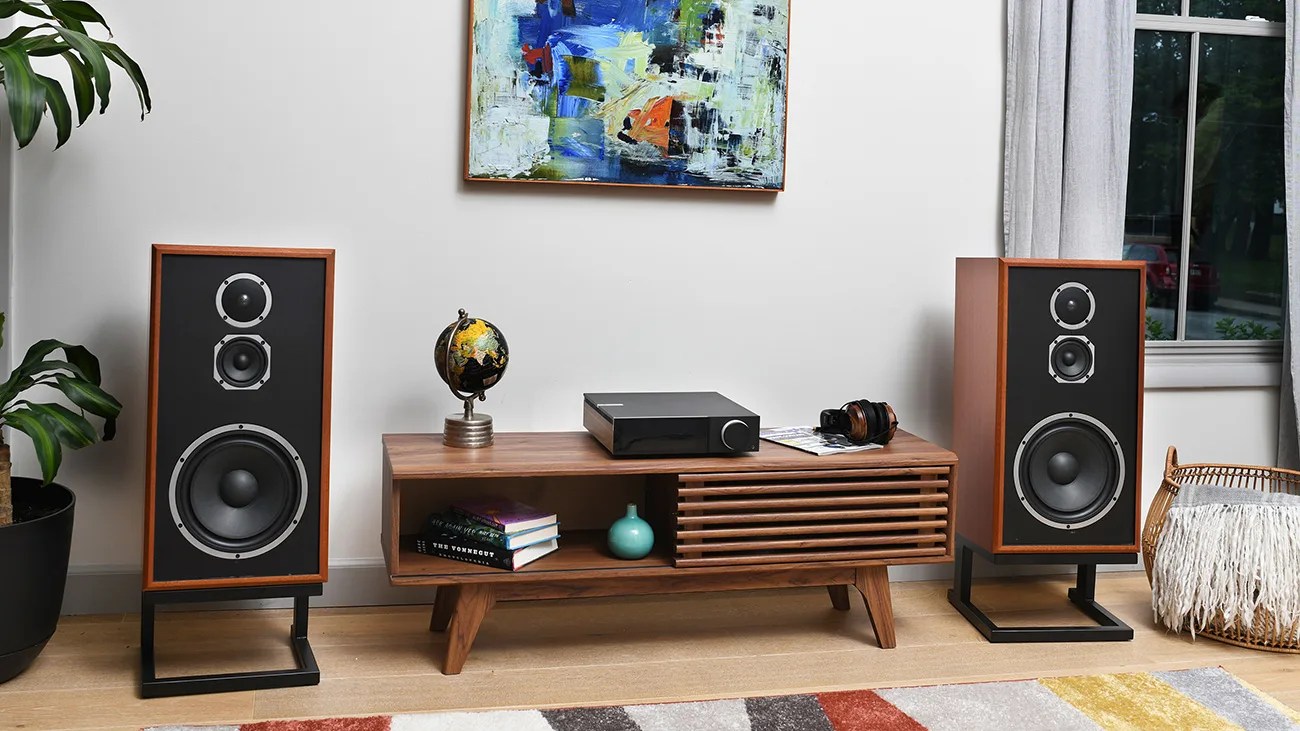 Pair of large wooden floor-standing speakers with black fronts on metal stands beside a mid-century modern wooden media console.