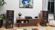 Pair of large wooden floor-standing speakers with black fronts on metal stands beside a mid-century modern wooden media console.