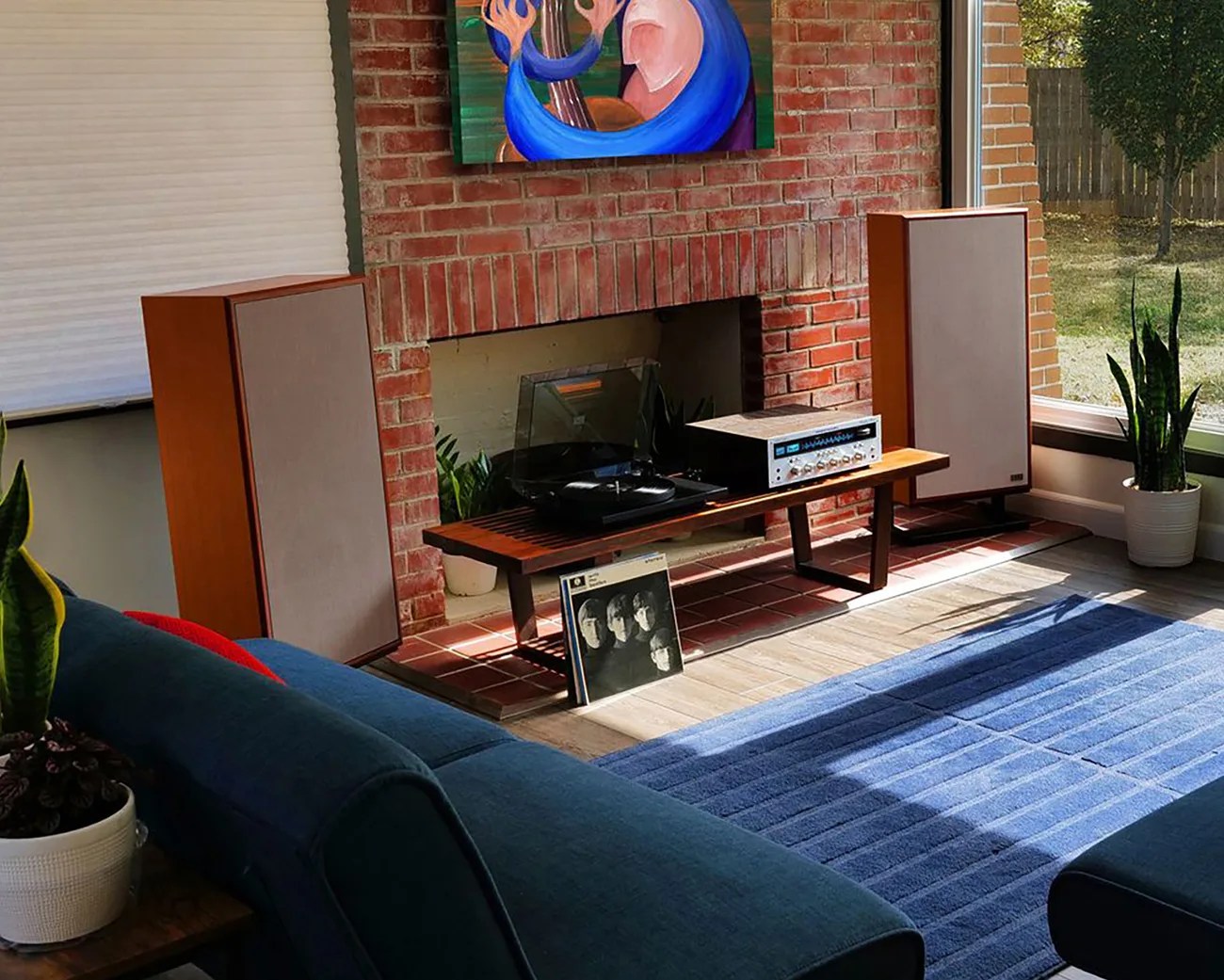 Mid-century living room with wooden speakers, turntable, amplifier on a wooden bench, blue rug, and brick fireplace.
