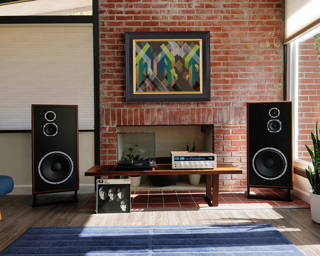 Pair of large vintage black and wood speakers flanking a wooden bench with a turntable, amplifier, and a Beatles vinyl in a room with brick fireplace.