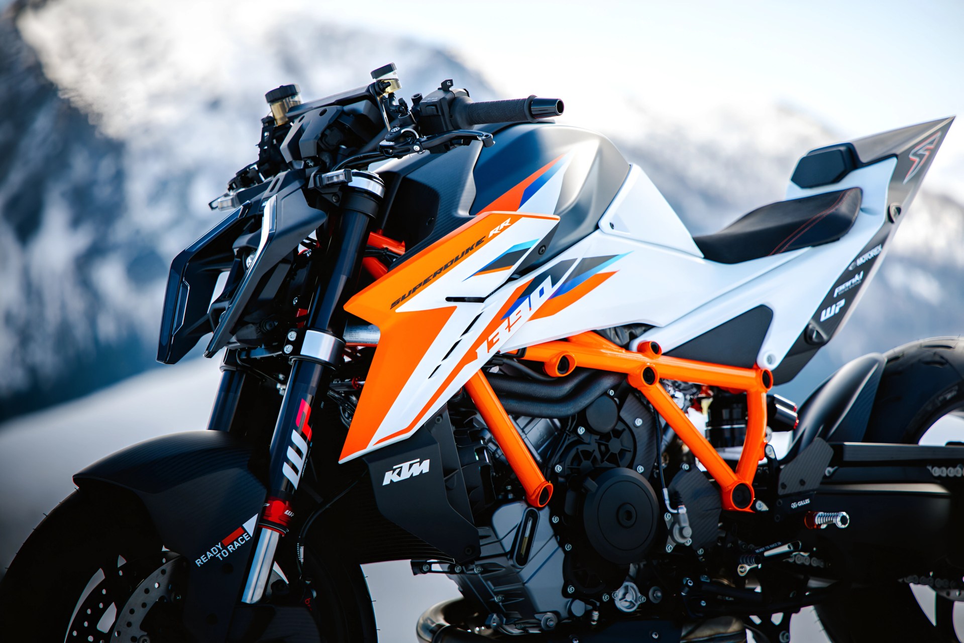 Close-up of a KTM 1290 Super Duke RR motorcycle with orange frame and white, orange, and blue body panels against a snowy mountain background.