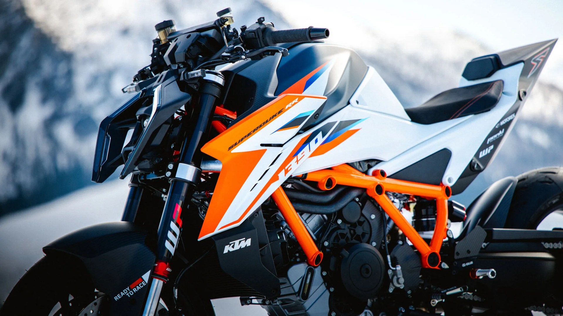 KTM Super Duke RR motorcycle with orange frame and white, black, and orange body panels against a snowy mountain background.