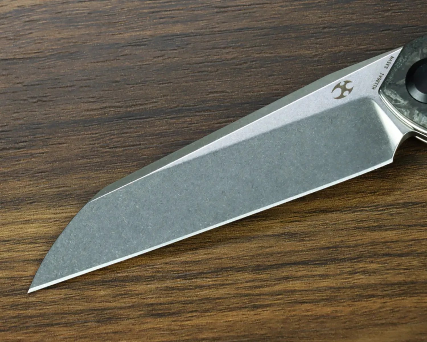 Knife blade with a stonewashed finish and a logo near the handle on a wooden surface.