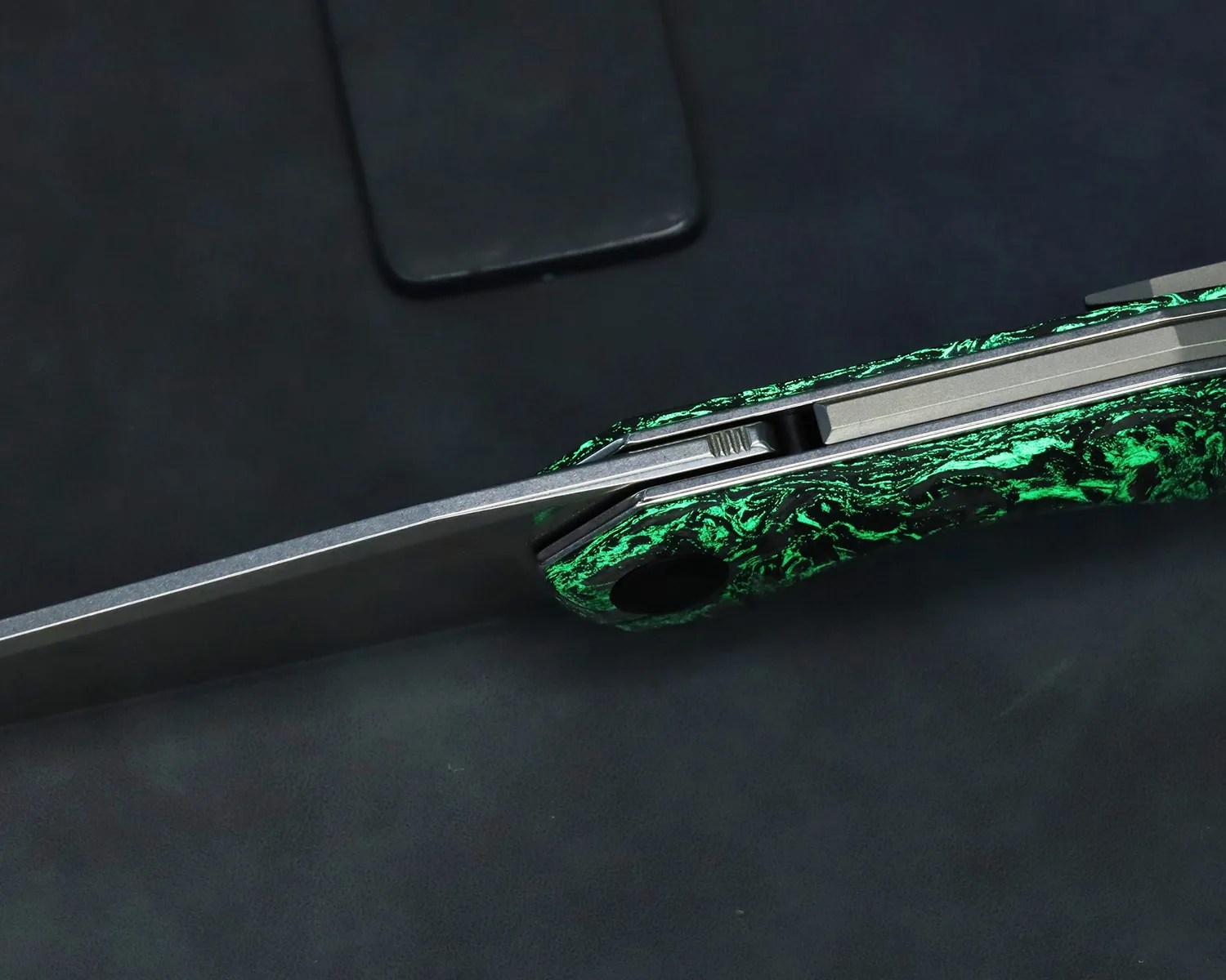 Close-up of a folding knife with a green and black marbled handle and a partially visible blade on a dark surface.