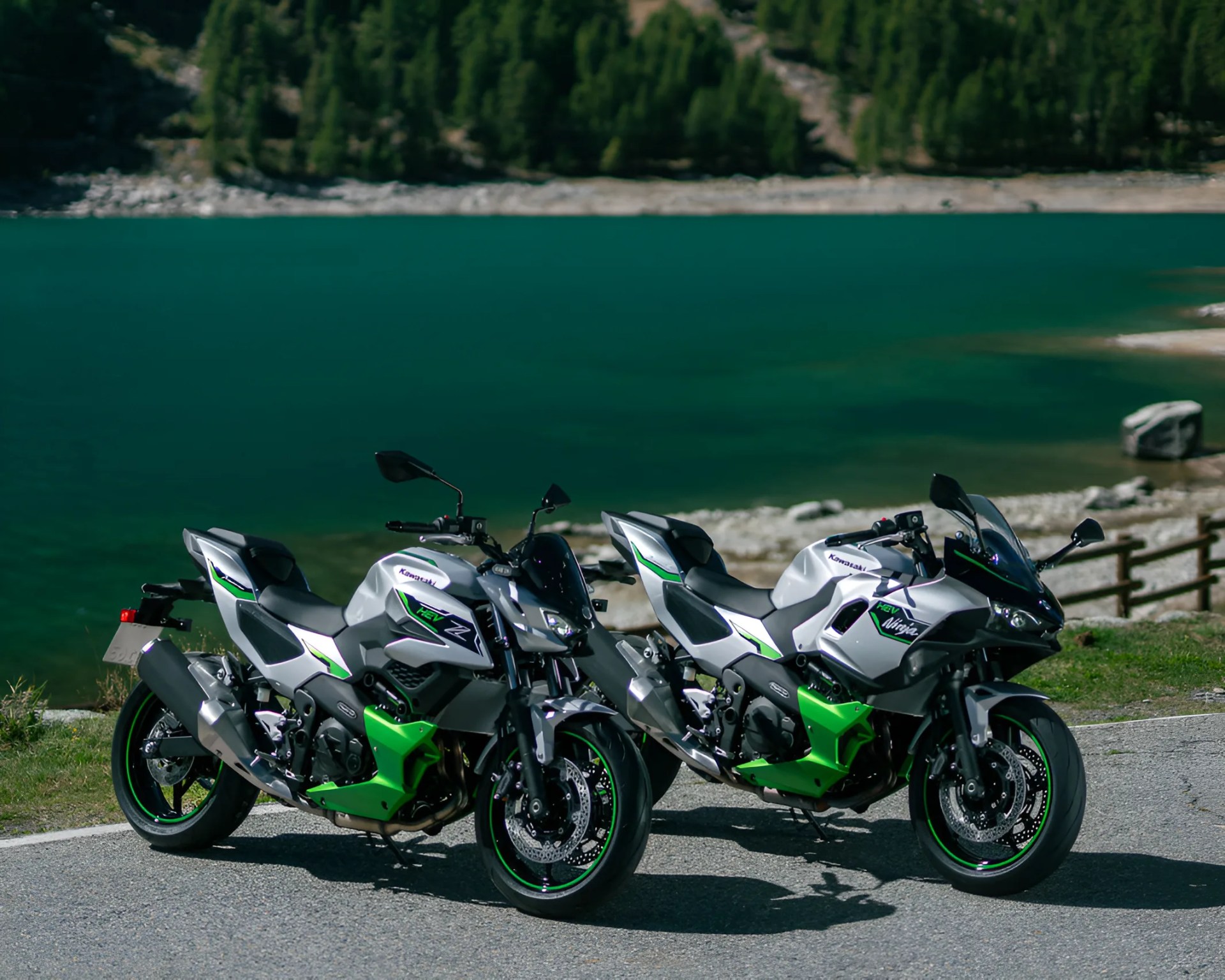 Two silver and green Kawasaki motorcycles parked on a lakeside road with trees in the background.