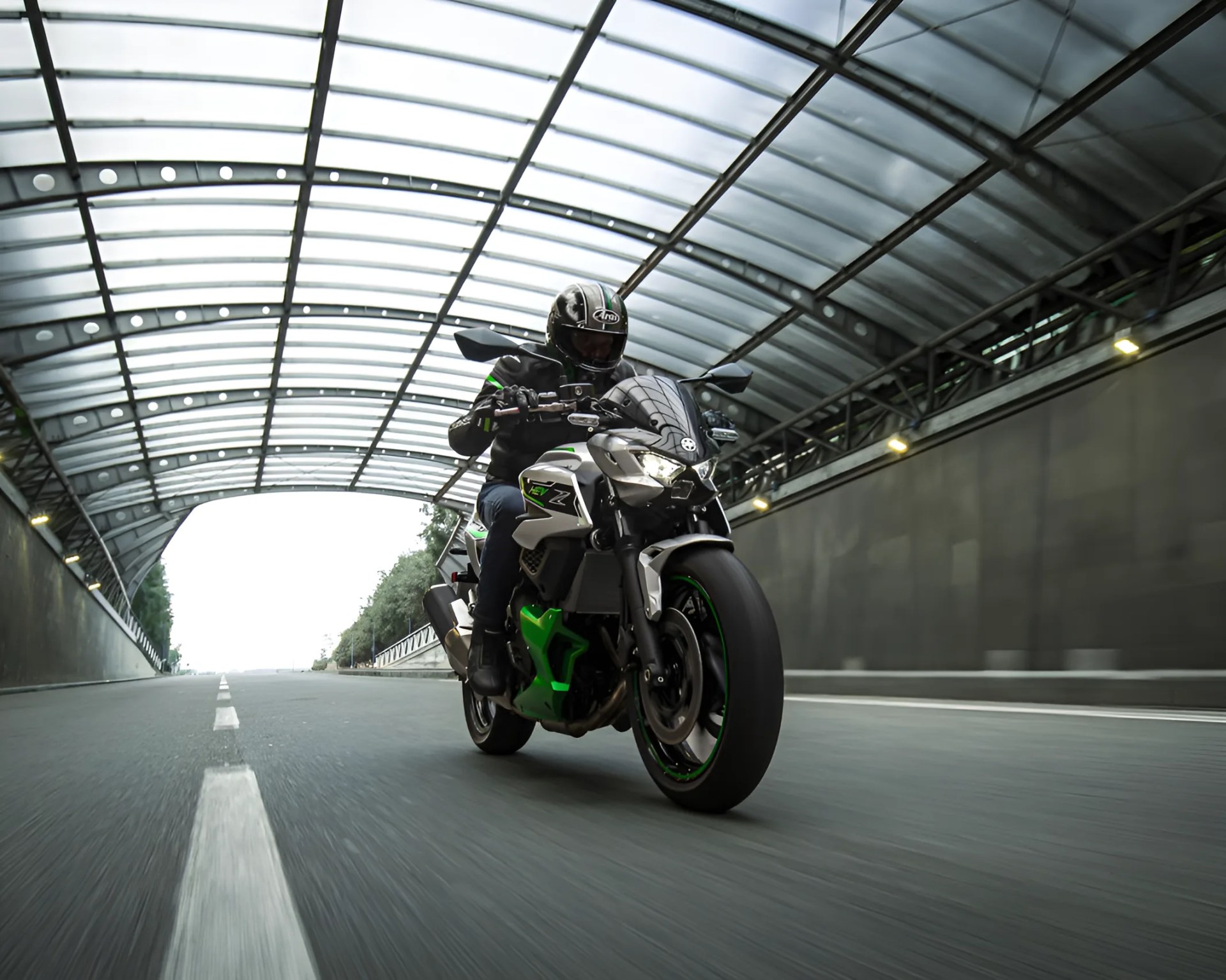 Sport motorcycle with green and silver accents ridden by a person wearing a black helmet and jacket in a tunnel with a glass roof.