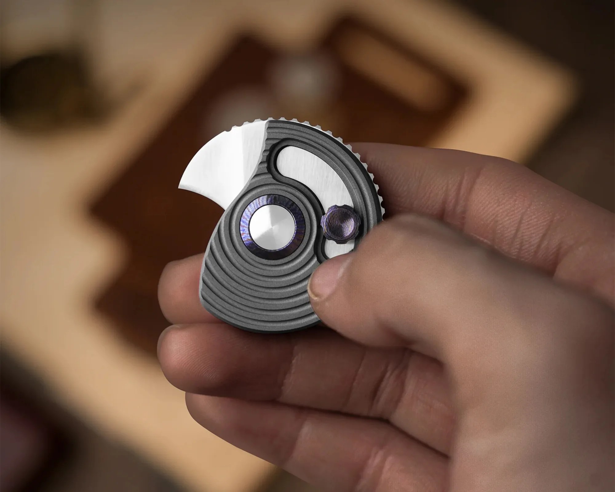 Hand holding a small, round, gray metal tool with a curved blade and a purple adjustment knob.