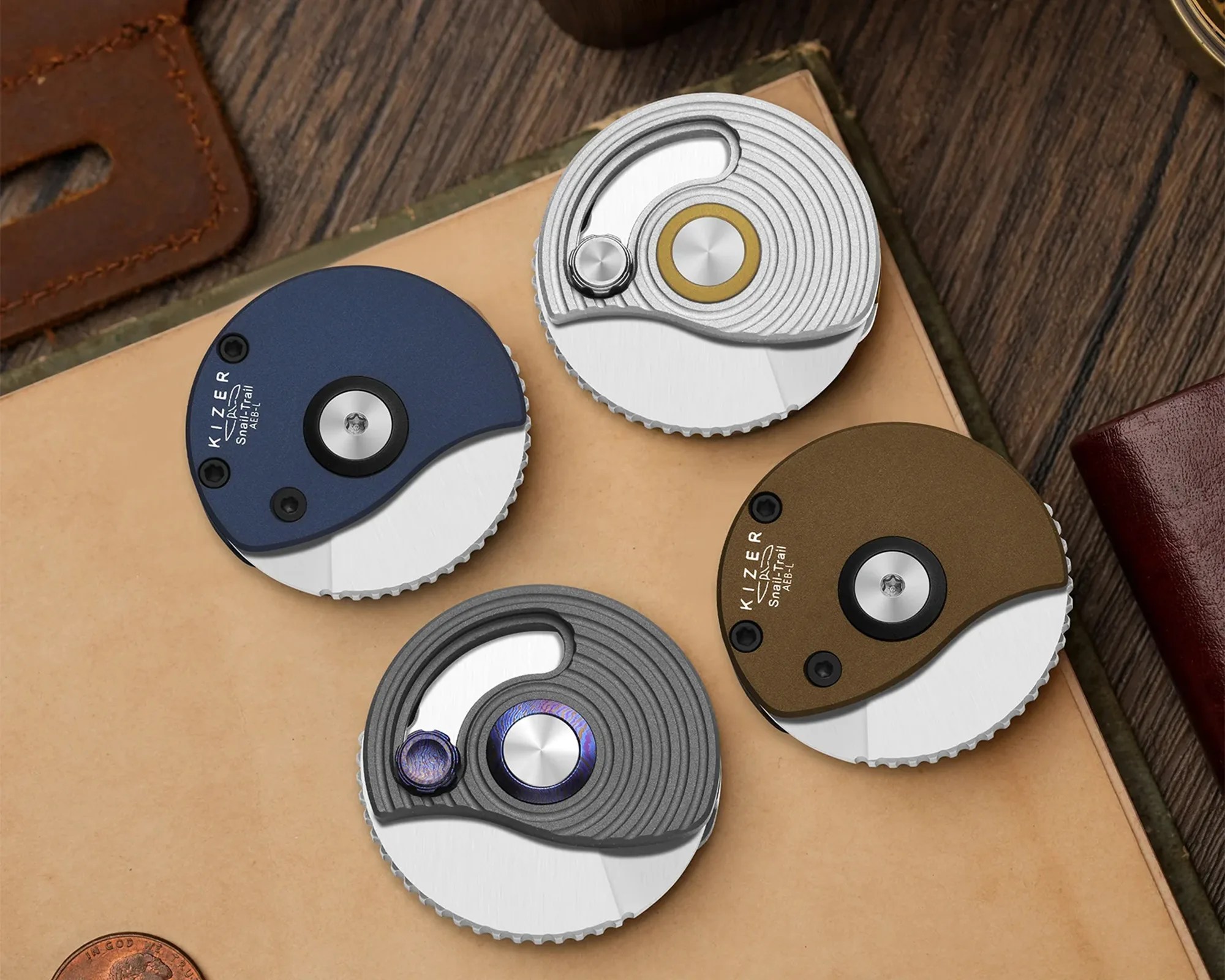 Four round folding knives with partially exposed blades in blue, silver, bronze, and gray finishes on a tan surface.