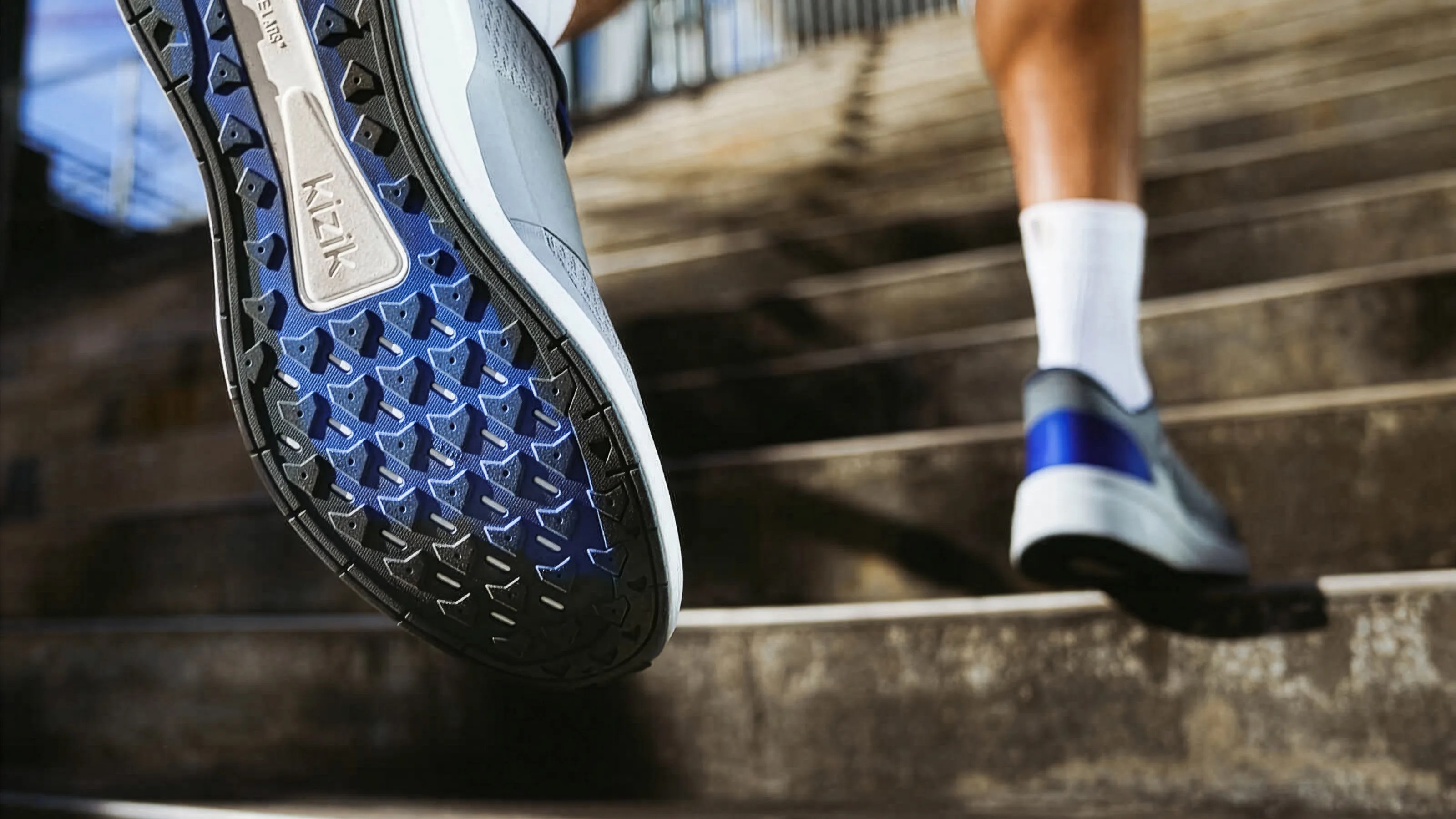 Close-up of a blue and black Kizik shoe sole with triangular treads on a person walking up stone stairs.