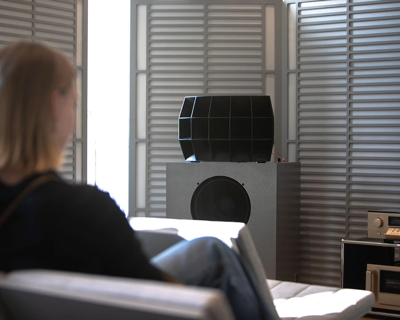 Person sitting on a chair facing a large gray speaker with a black horn on top in a room with gray acoustic panels.