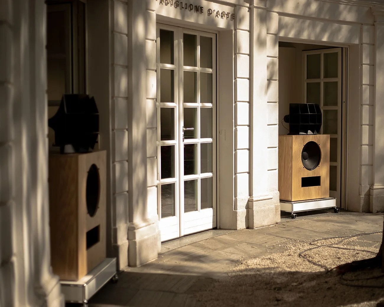 Pair of large wooden speakers with black horn tweeters placed outside white double glass doors in a sunlit courtyard.