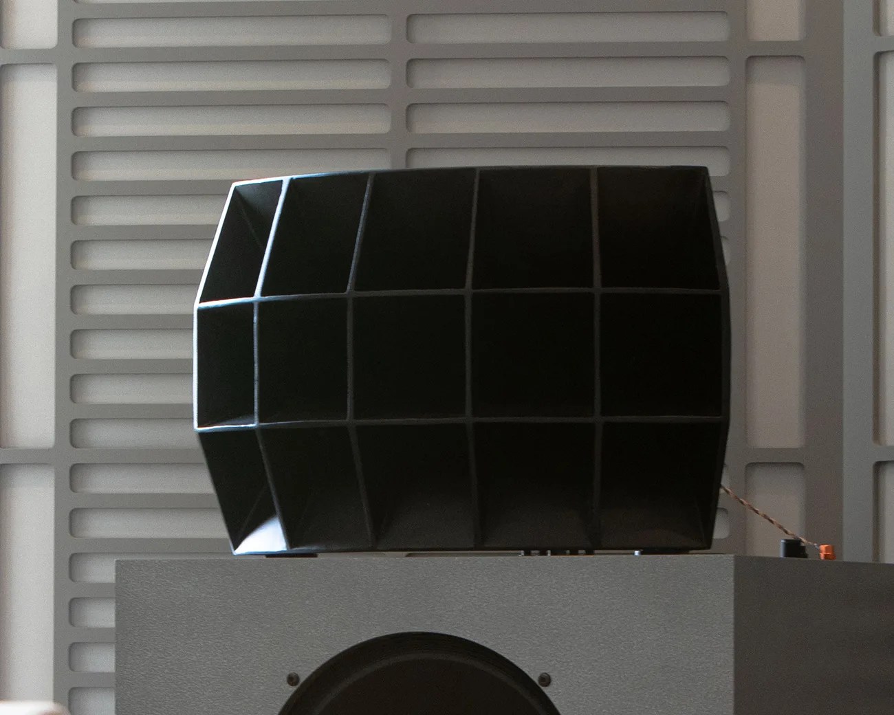 Black multi-chambered speaker horn mounted on a gray speaker cabinet against a gray wall panel.