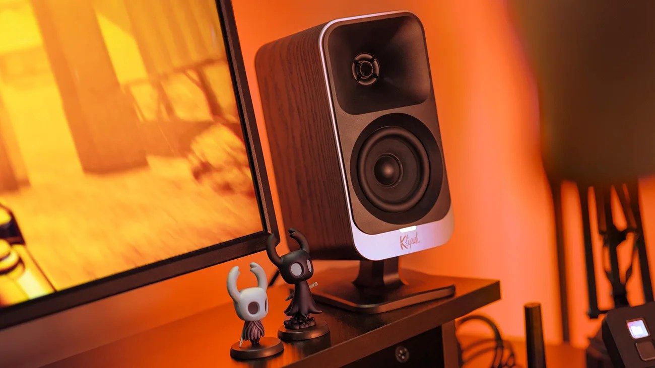 Wood-finished Klipsch speaker on a stand next to two small black and white figurines on a desk.