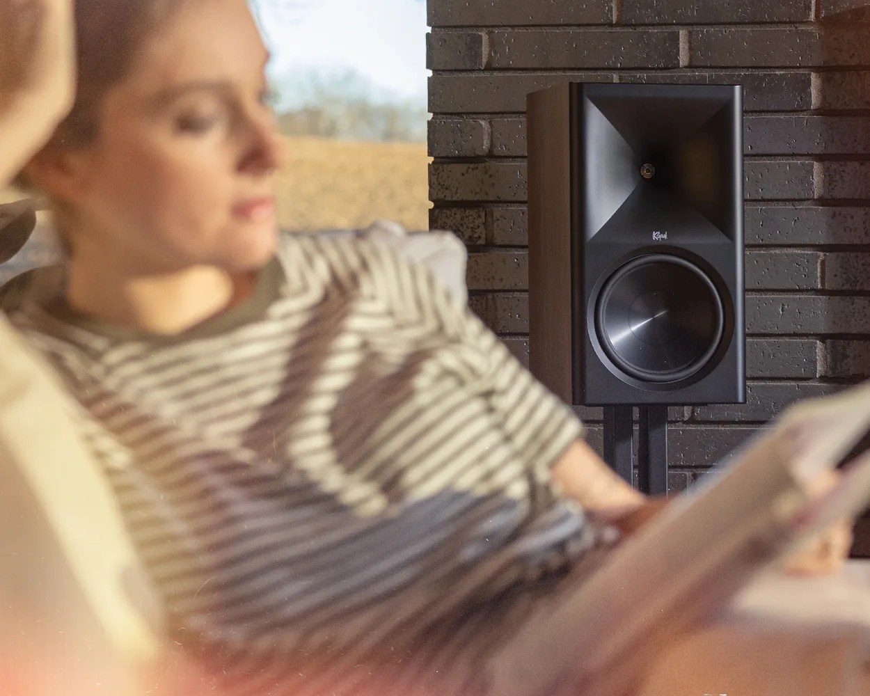 Black Klipsch speaker on a stand against a dark brick wall with a person reading in the foreground.