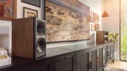 Pair of bookshelf speakers with wood veneer sides and black front panels on a long black cabinet under a wall-mounted TV.