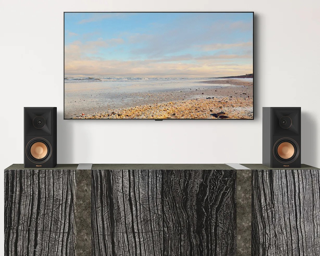 Two black Klipsch speakers with copper cones on a textured black and gray cabinet below a wall-mounted flat-screen TV showing a beach scene.