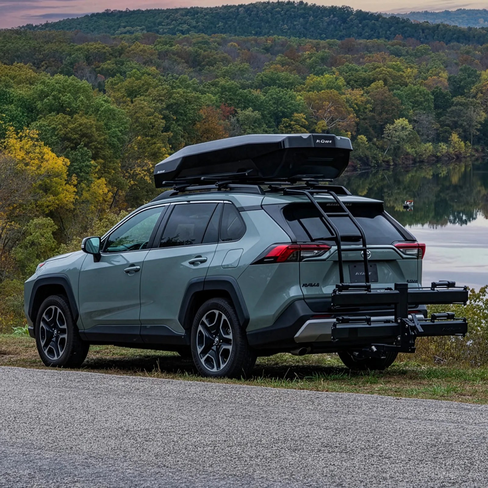 Green Toyota RAV4 with black roof cargo box and rear bike rack parked near a lake with forested hills in the background.