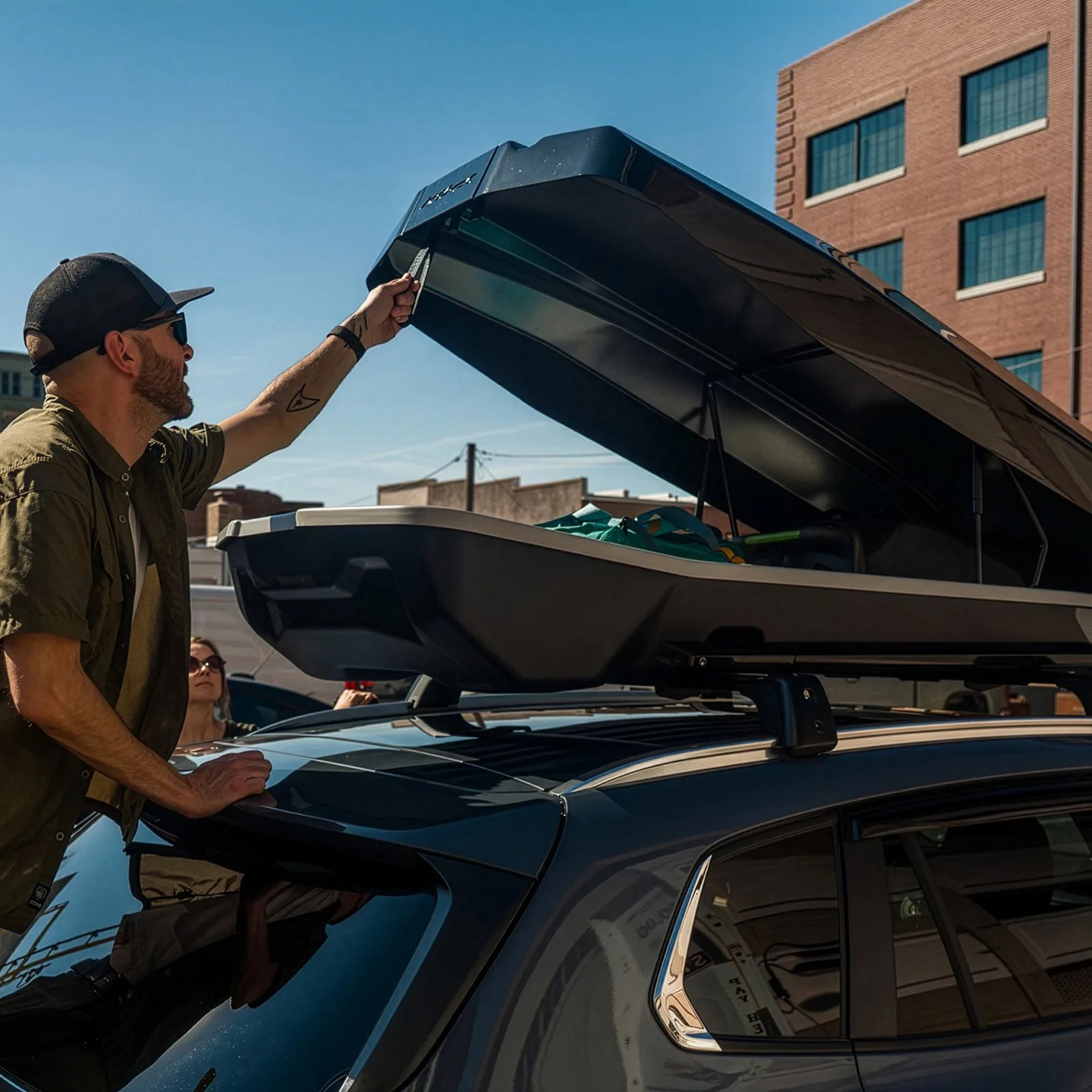 Man in a cap opening a black rooftop cargo box mounted on a dark SUV in an urban setting.