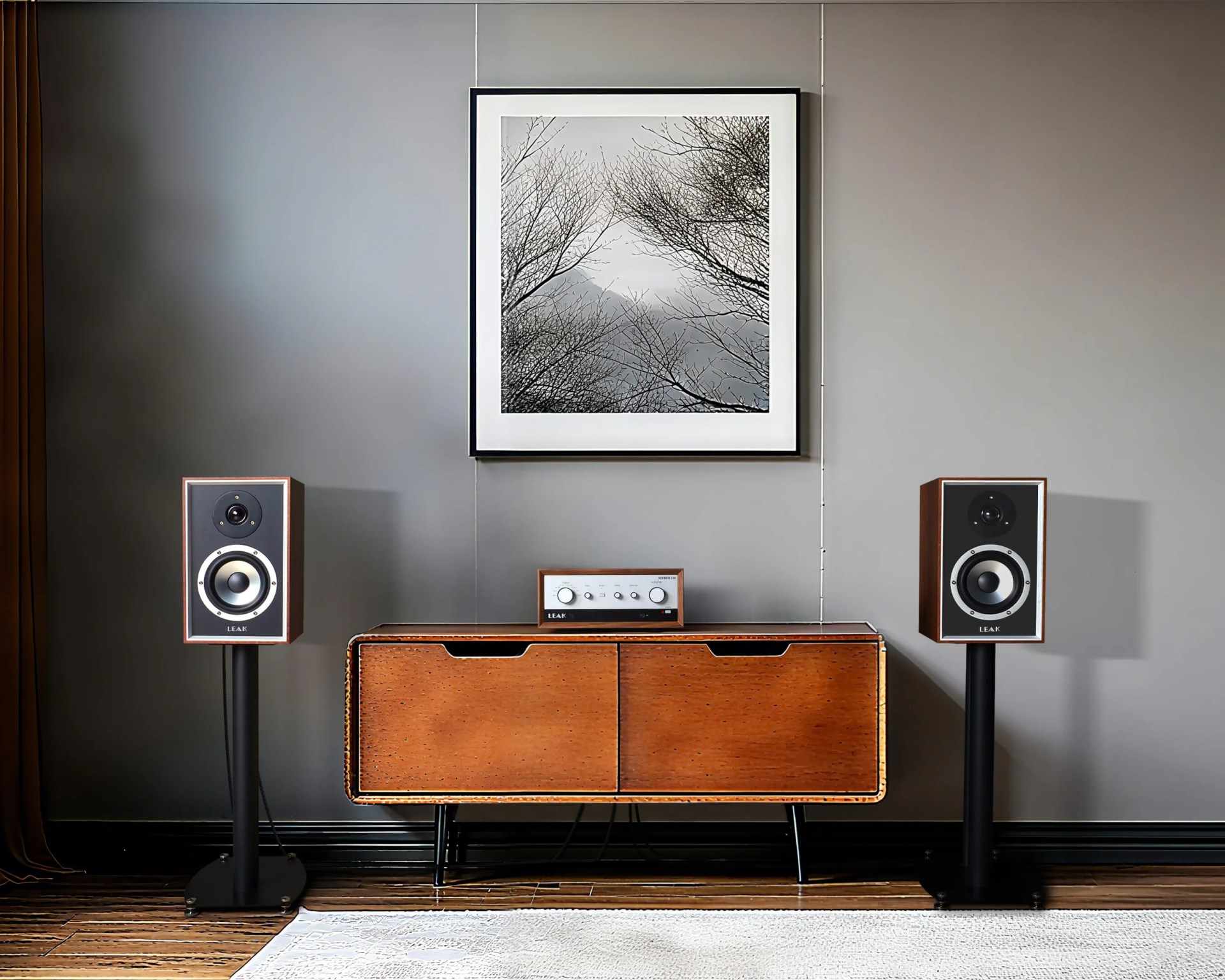 Pair of wooden bookshelf speakers on black stands flanking a wooden cabinet with a stereo amplifier on top, under a framed black-and-white tree artwork.