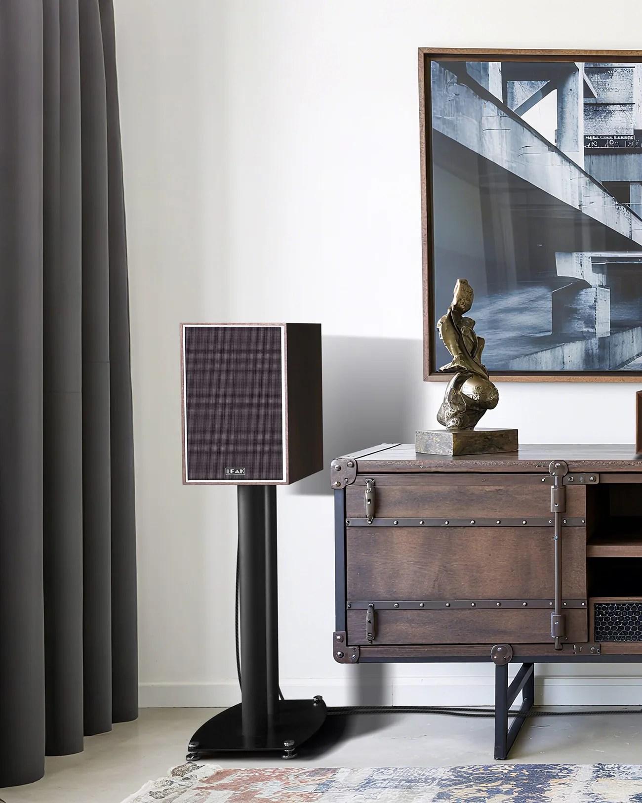 Brown LEAK speaker on a black stand next to a wooden cabinet with a bronze sculpture and framed abstract photo above.