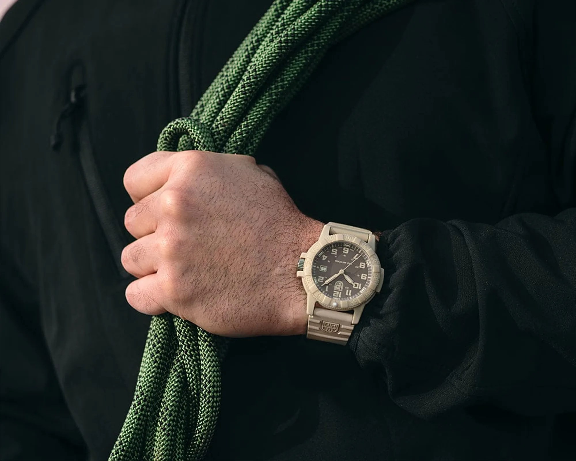 Beige tactical wristwatch with black dial and white numerals worn on a wrist holding green climbing rope.