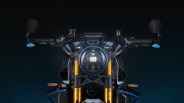 Front view of a motorcycle headlight with gold front forks, round LED light, and black handlebars with blue accents.