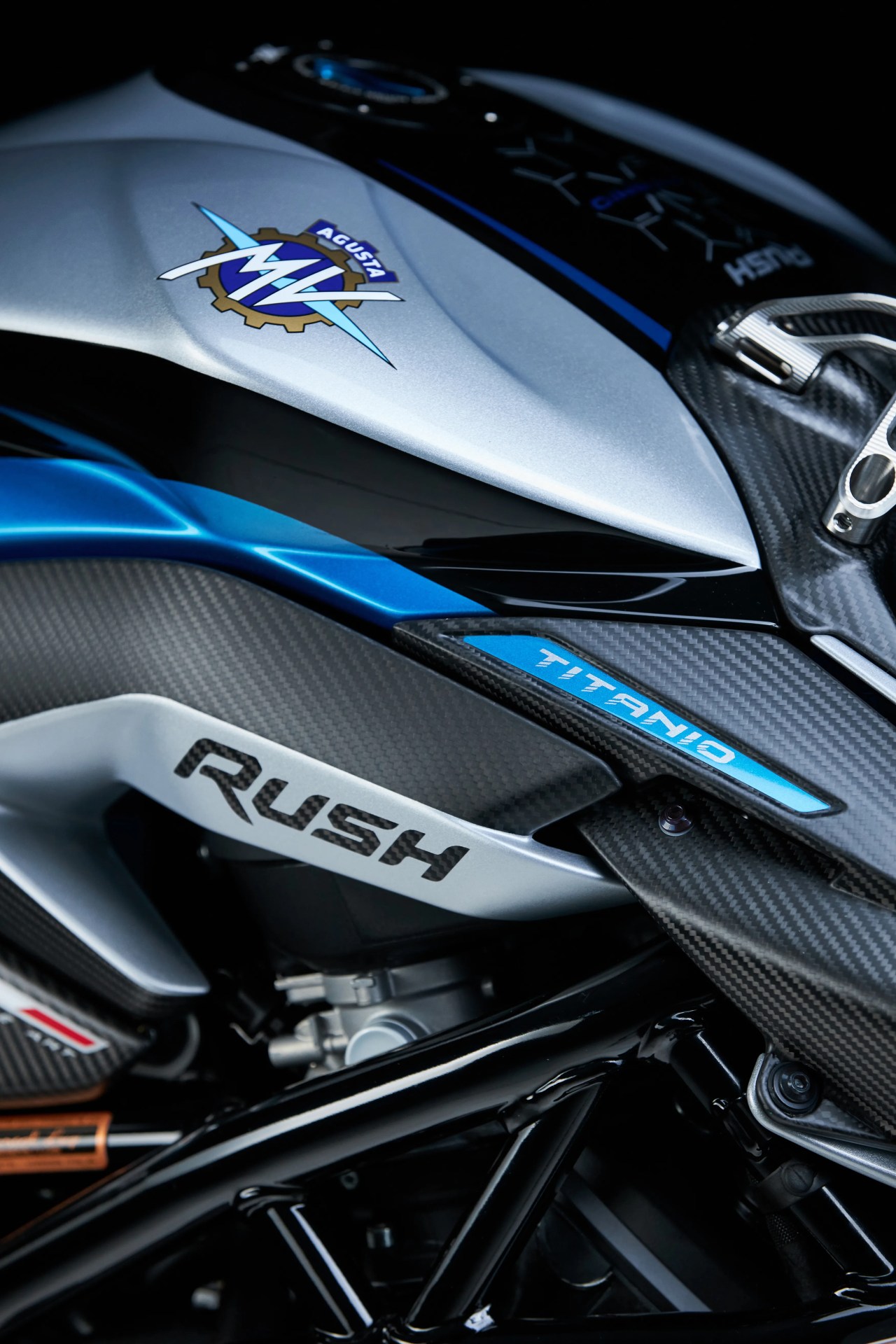 Close-up of a silver and blue MV Agusta motorcycle fuel tank with "Rush" and "Titanio" labels and carbon fiber detailing.