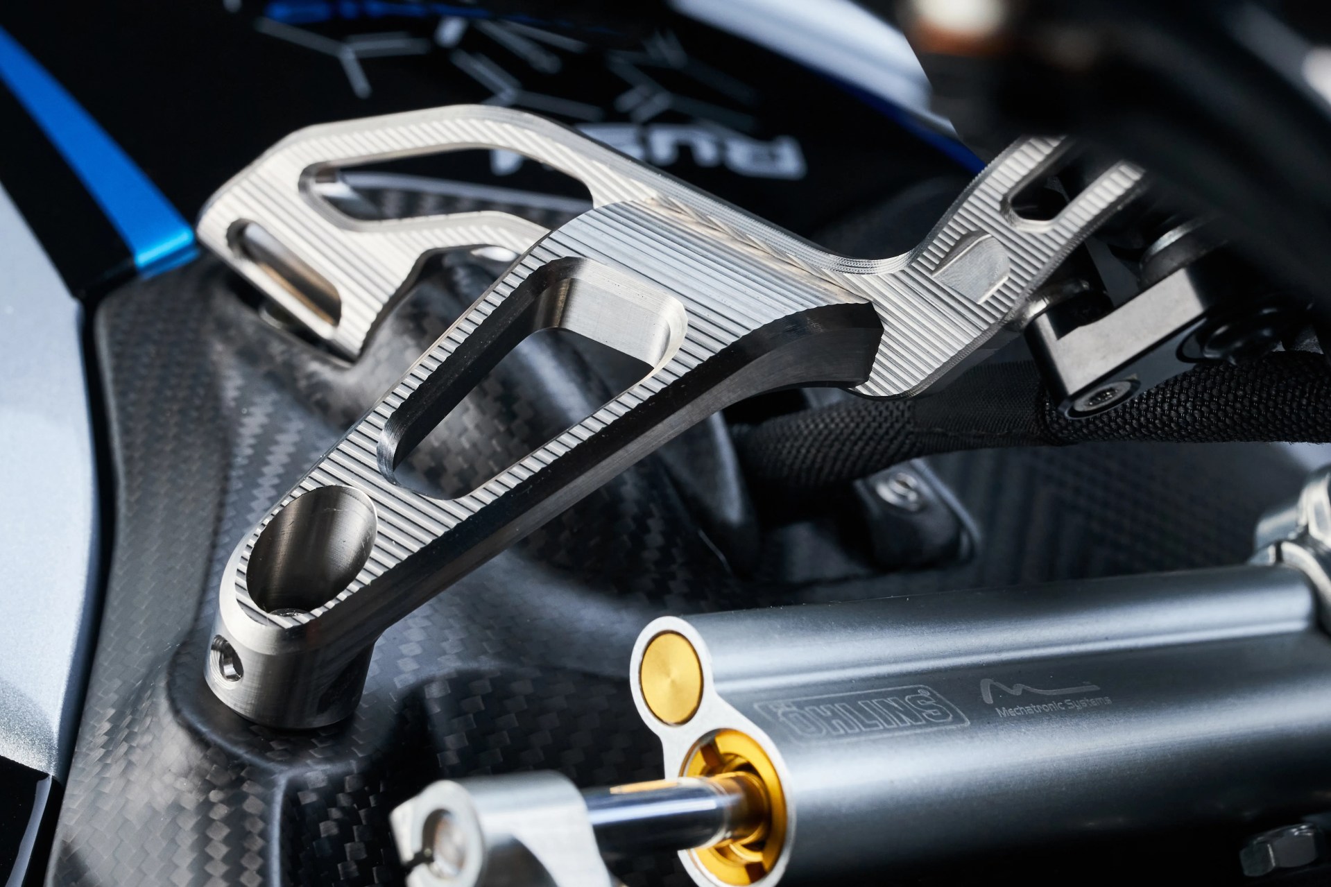 Close-up of a silver metal motorcycle part mounted on a carbon fiber surface next to a gray Öhlins suspension component.