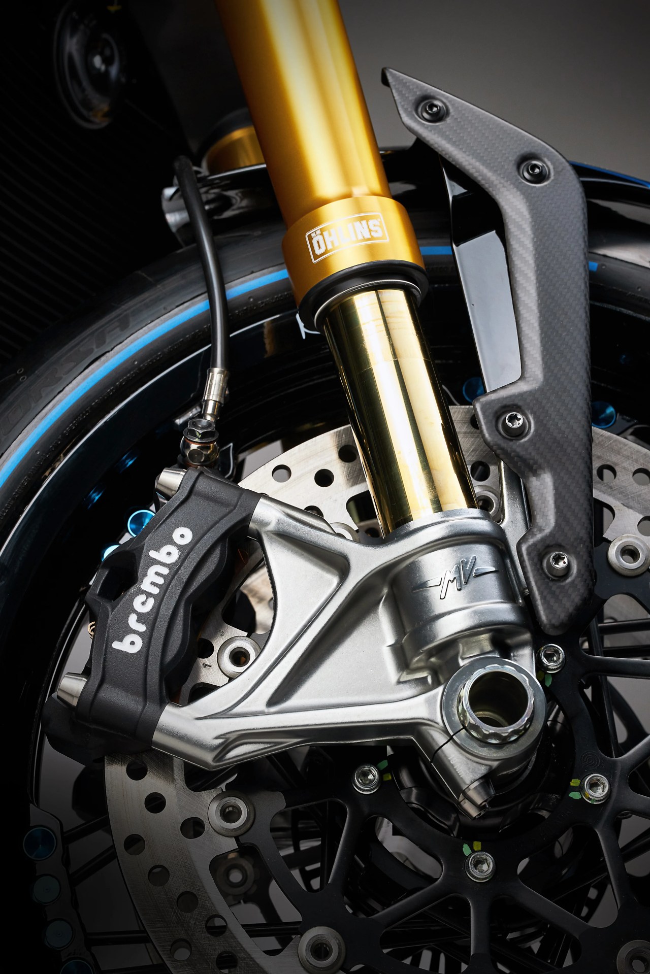 Close-up of a motorcycle front suspension with gold Öhlins fork, Brembo brake caliper, and carbon fiber guard.