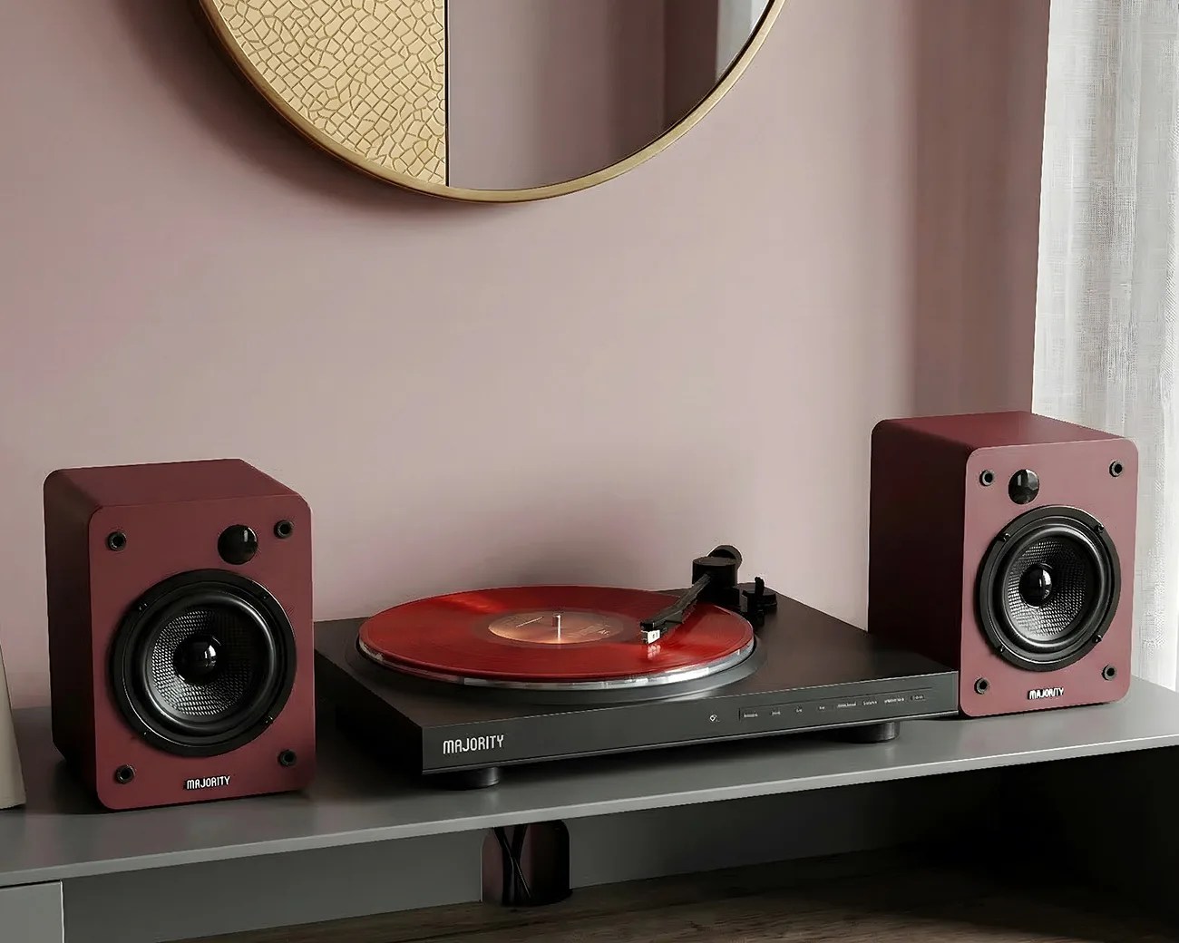 Black turntable with red vinyl and matching maroon speakers on a gray shelf under a round mirror.