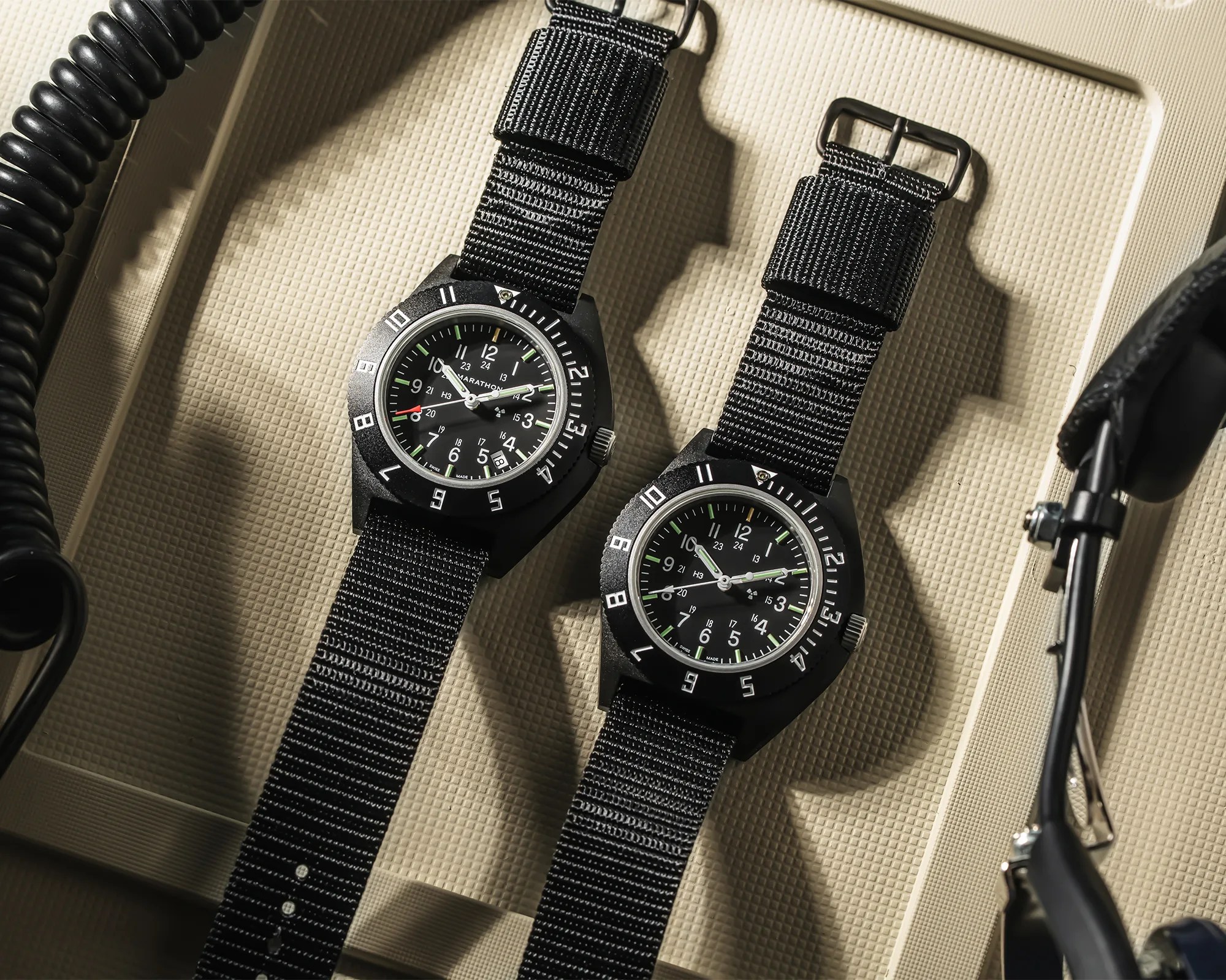 Two black Marathon wristwatches with black nylon straps on a textured beige surface.